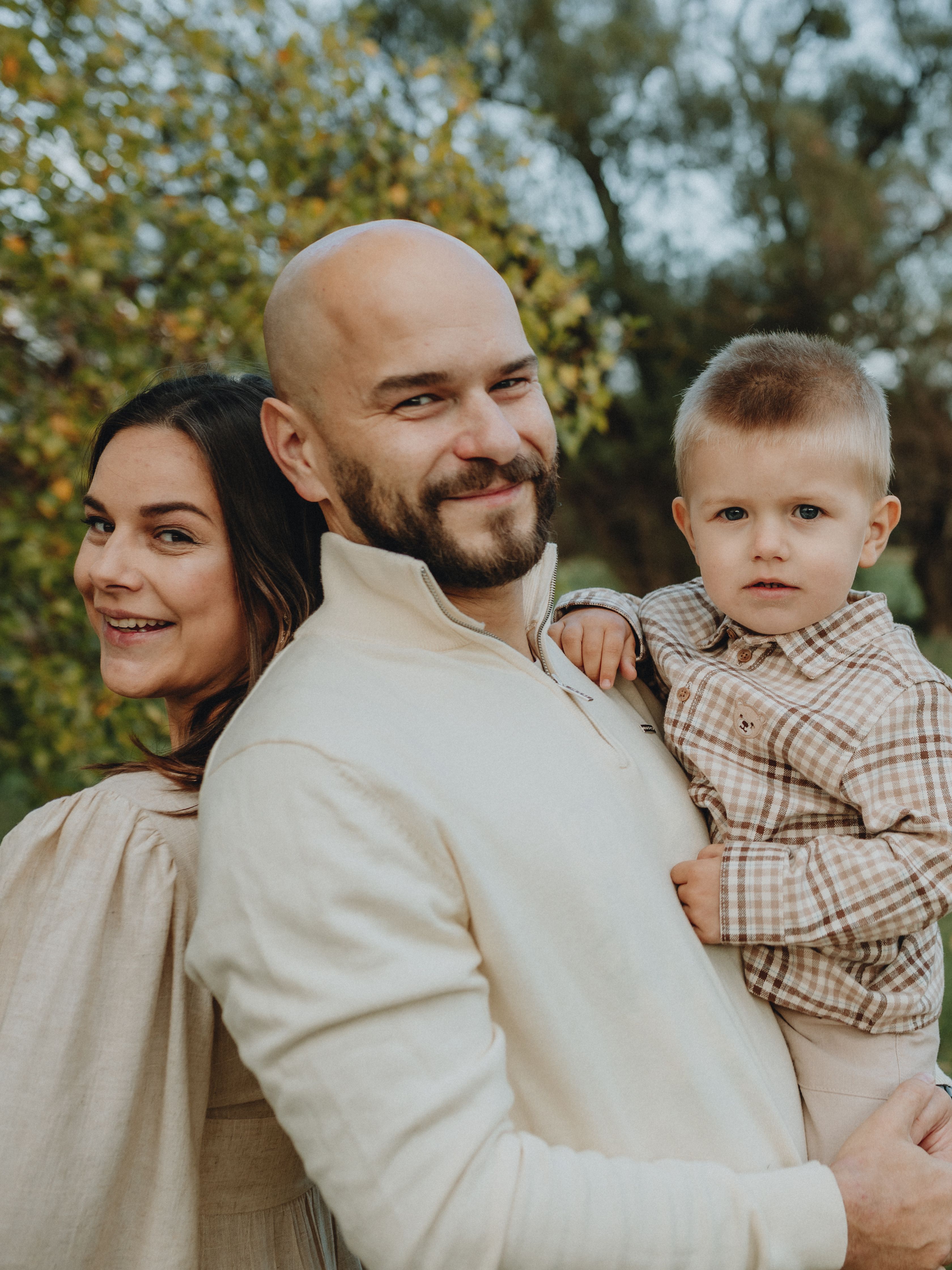 A family of three smiling together outdoors among autumn trees