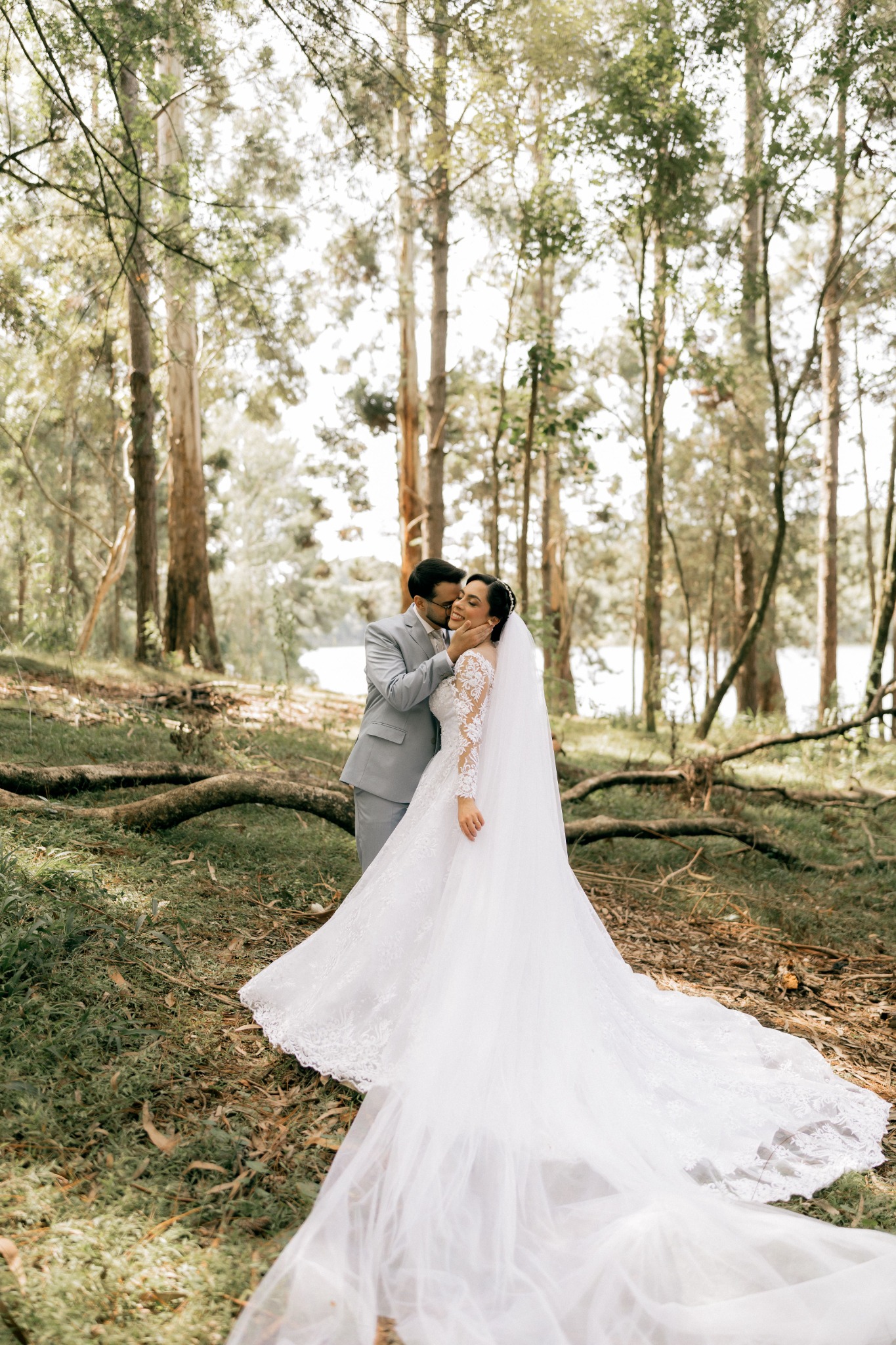 A groom kissing his smiling bride among sunlit trees, with her train spread across the grass