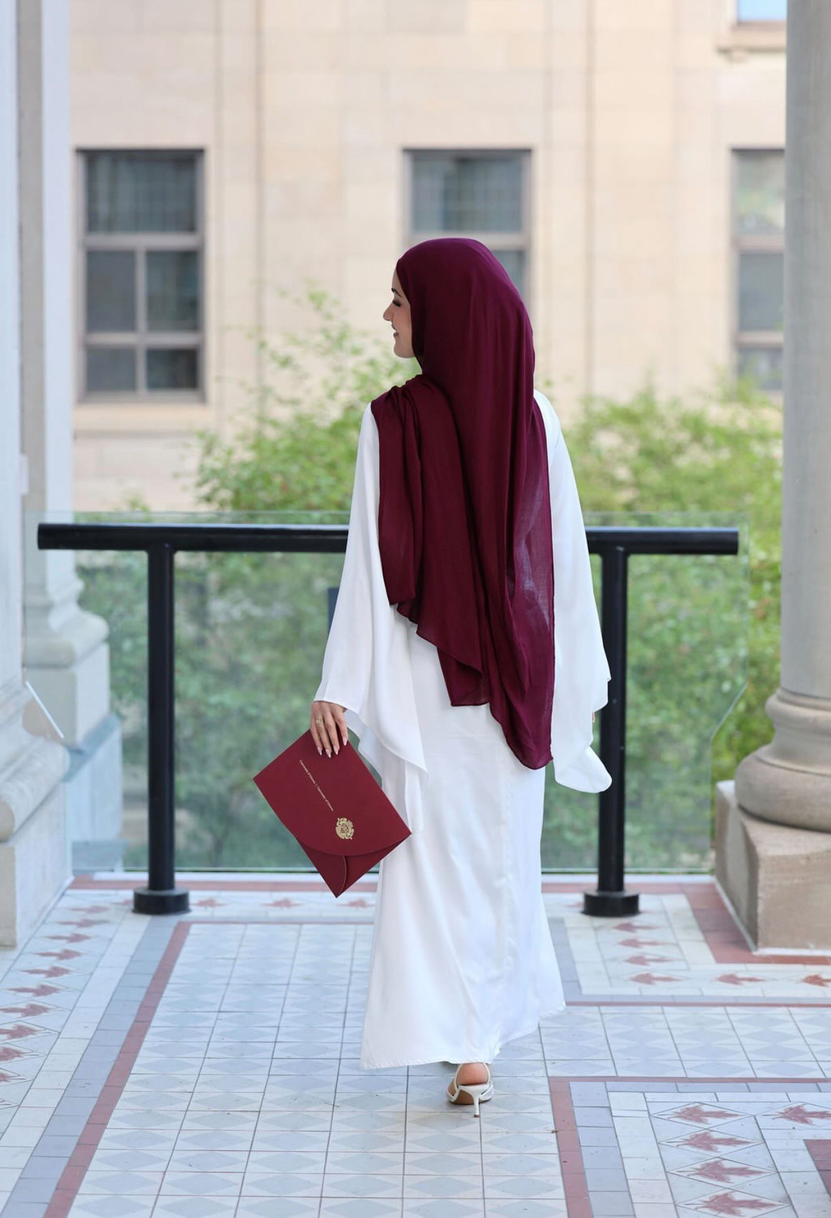 Graduate in a white outfit and burgundy headscarf holding a diploma folder